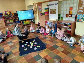 Dzieci siedzą w kole. W środku klęczy nauczycielka, która przed sobą ma rozłożony granatowy materiał z przyczepionymi cyframi i kolorowymi figurami geometrycznym