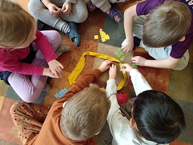 6 dzieci siedzi w kole. W środku leży żółta szarfa, w której znajdują się kolorowe figury geometryczne. Dzieci wyciągają figury i segregują je ze względu na kolor.