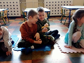 Dwóch chłopców siedzi na macie i trzyma w dłoniach zabawkowego ślimaka. Chłopcy śmieją się do siebie