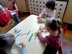Dzieci siedzą przy stoliku i ozdabiają papierowe pudełka. Na stoliku leżą różne kolory flamastrów.