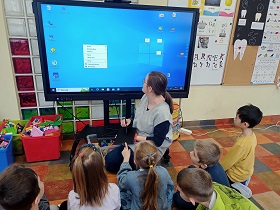 Dzieci siedzą przed monitorem i patrzą na panią, która pokazuje im jak działa tablet graficzny, który trzyma na kolanach. Na monitorze wyświetla się niebieski ekran komputerowy z aplikacjami.