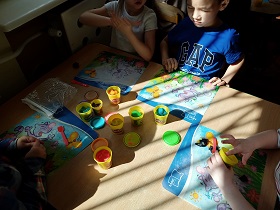 Czworo dzieci siedzi przy stoliku i bawi się ciastoliną ugniatając ją na plastikowych tackach. Na stoliku znajdują się również pojemniki z ciastoliną.