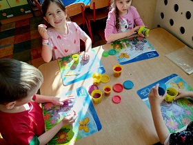 Czwórka dzieci siedzi przy stoliku i bawi się ciastoliną ugniatając ją na plastikowych tackach. Na stoliku znajdują się również pojemniki z ciastoliną.