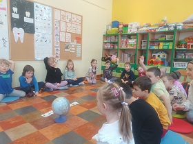Dzieci siedzą na podłodze, na kolorowych poduszkach i się zgłaszają. Przed nimi na środku leży globus i napis globus z papierowych karteczek. 