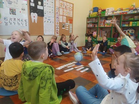 Dzieci siedzą na podłodze i wskazują swoją jedną dłonią pięć palców podnosząc rękę do góry. Na środku kółka leży globus oraz napisy.