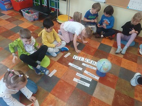 Dzieci siedzą na kolorowych poduszkach na podłodze i mają przed sobą prostokątne małe karteczki. Obok nich leży globus oraz napisy. 