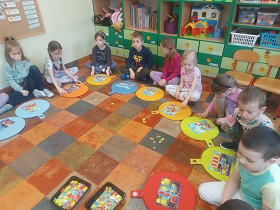 Dzieci siedzą za kolorowymi poduszkami na podłodze i mają przed sobą prostokątne obrazki, na których ułożone są kolorowe figury geometryczne. Obok nich leżą pudełka z figurami geometrycznymi.