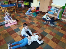 Na zdjęciu widać 8 dzieci- 4 dziewczynki i 4 chłopców. Dzieci dobrane są w pary. Jedno dziecko z pary leży na podłodze, na macie a drugie klęczy obok niego i wykonuje masaż pleców. 