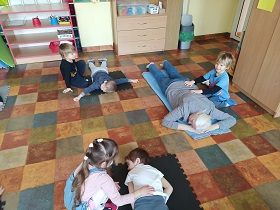 Na zdjęciu widać 4 chłopców, dziewczynkę i nauczycielkę. Nauczycielka leży na kocu na podłodze i jest masowana przez chłopca, który klęczy obok niej. 4 dzieci dobrane jest w pary. Jedna osoba z pary leży na macie, na podłodze a druga klęczy obok i wykonuje masaż pleców. 