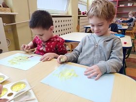 Dwóch chłopców siedzi przy stoliku. Przed nimi leżą błękitne kartki z namalowanymi żółtymi kurczaczkami. Jeden z chłopców trzyma w ręku patyczek z czerwoną farbą a drugi widelec zamoczony w żółtej farbie. 