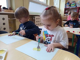 Chłopiec i dziewczynka siedzą przy stoliku. Na błękitnych kartkach malują żółte kurczaczki przy pomocy farby i widelca.