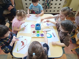 Chłopiec i 5 dziewczynek siedzą przy stoliku. Przed sobą mają kartki na których malują portret mamy. Pośrodku na stole stoi pojemnik z farbami oraz kubek z wodą. 
