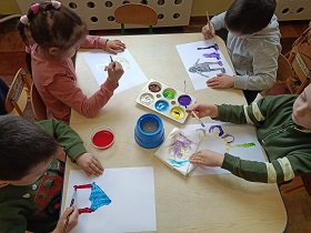 3 chłopców i dziewczynka siedzą przy stoliku. Przed sobą mają kartki na których malują portret mamy. Pośrodku na stole stoi pojemnik z farbami oraz kubek z wodą. 