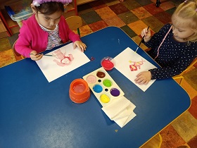 2 dziewczynki siedzą przy stoliku. Przed sobą mają kartki na których malują portret mamy. Pośrodku na stole stoi pojemnik z farbami oraz kubek z wodą. 
