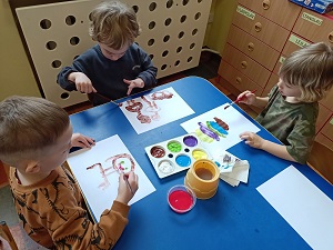 3 chłopców siedzi przy stoliku. Przed sobą mają kartki na których malują portret mamy. Pośrodku na stole stoi pojemnik z farbami oraz kubek z wodą. 
