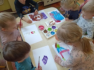3 chłopców i 3 dziewczynki siedzą przy stoliku. Przed sobą mają kartki na których malują portret mamy. Pośrodku na stole stoi pojemnik z farbami oraz kubek z wodą. 