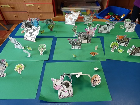 Na stole leży 8 przestrzennych prac dzieci. Na zielonych kartkach przyklejone są pokolorowane i wycięte krowy, drzewa oraz krzaki. 