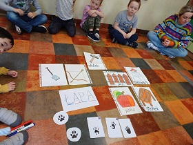 Dzieci siedzą w kole. Po środku leżą obrazki z napisem ŁAPA, rysunki łopaty, jabłka, łuku, młotka, bałwana, stołka oraz ślady łap różnych zwierząt. 