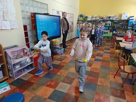 Dzieci ustawione są w dwóch rzędach, podzielone na dwie drużyny. Dzieci biorą udział w zawodach w jak najszybszym przejściu z małą piłeczką na łyżce. Na zdjęciu konkuruje ze sobą chłopiec ubrany na szaro i dziewczynka w beżowej bluzie.