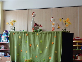Wiosenny teatrzyk kukiełkowy jest ozdobiony żonkilami i tulipanami. Zdjęcie przedstawia scenkę odgrywaną przez kukiełkę kury i koguta.