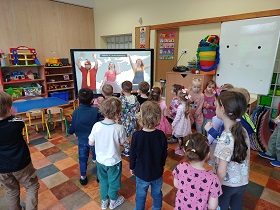 Dzieci stoją przed monitorem i wykonują ruchy, które pokazują im trzy osoby z filmiku.