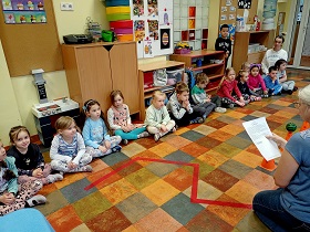 Dzieci siedzą w rzędzie na podłodze. Pani w niebieskiej bluzce czyta im tekst. 
