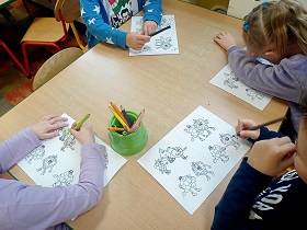 Czworo dzieci siedzi przy stole i koloruje obrazki. Na środku stołu stoi pudełko z kredkami. 