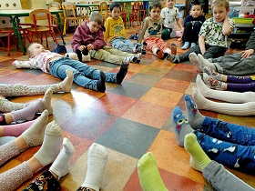 Dzieci siedzą w kółeczku. Mają wyciągnięte nogi bez butów, w samych skarpetkach. 