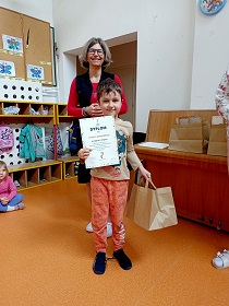 Chłopiec stoi obok pani i trzyma w dłoniach dyplom oraz papierową torbę. 