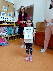 Dziewczynka stoi obok dwóch pań i trzyma w dłoniach dyplom.