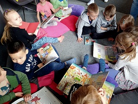 Dzieci siedzą w kółeczku na poduszkach i oglądają książki dla dzieci. 