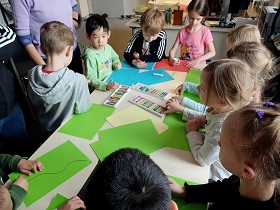 Dzieci stoją wokół stołu. Na stole leżą kolorowe kartki i pastele. 