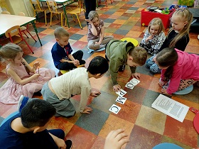 Na podłodze chłopiec w zielonej bluzie układa słowo pomoc z kartoników z literami. Reszta dzieci się temu przygląda.