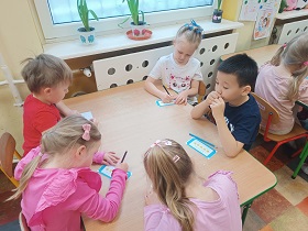 Piątka dzieci siedzi przy stole i kredkami zaznacza coś na kartce w niebieskiej ramce.