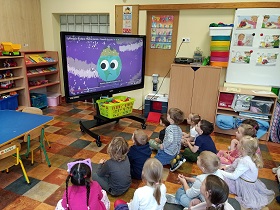 Dzieci siedzą w sali na podłodze. Przed nimi znajduje się tablica multimedialna z wyświetlonym teledyskiem do piosenki "Nasza planeta". 
