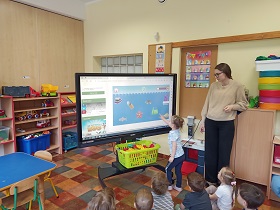 Dzieci znajdują się w sali i siedzą na podłodze przed tablicą multimedialną na której wyświetlona jest grą przedstawiające zaśmiecony ocean. Obok ekranu stoi nauczycielka. Dziewczynka stoi przy ekranie i palcem wskazuje na ilustracje przedstawiająca torebkę foliową.