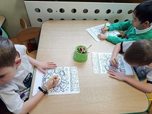 Przy stole siedzi 3 chłopców. Każdy z nich ma przed sobą kolorowankę przedstawiająca ziemię, uśmiechającą się dzieci i napis "kochajmy zieleń, pielęgnujmy ja każdego dnia". Na środku stołu stoi pudełko z kredkami. 