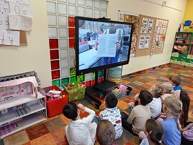 Dzieci siedzą przed monitorem edukacyjnym i oglądają proces powstawania papieru. Na ekranie widać fabrykę i osobę pokazującą książkę.