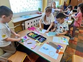 Dzieci siedzą przy stolikach i wycinają materiały w różnych kolorach i o różnych teksturach oraz wzorach. Na stolikach rozłożone są również kredki.