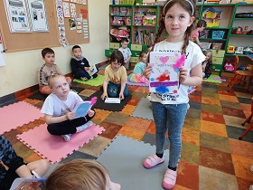 Dzieci siedzą na matach ze swoimi książkami. Dziewczynka w warkoczykach prezentuje na stojąco swoją książkę.