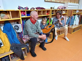 Chłopiec siedzi na ławce i trzyma gitarę klasyczną z pomocą dwóch Panów.