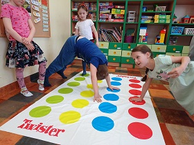 W Sali rozłożona jest mata do gry w Twister. Chłopiec ubrany na niebiesko trzyma ręce obie na macie. Dziewczynka w zielonej sukience trzyma ręce na czerwonym polu. Dwie pozostałe dziewczynki czekają na swoją kolej. 