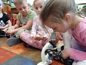 Dziewczynka w różowej sukience siedzi na podłodze i ogląda próbkę kurzu pod mikroskopem. 