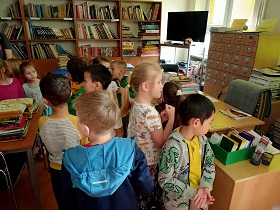 Dzieci znajdują się w bibliotece szkolnej i przyglądają się temu co się w niej znajduję. Z tyłu za dziećmi stoją regały z książkami. 