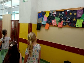 Trzy dziewczynki spacerują korytarzem szkolnym i podziwiają wiszące na tablicy prace plastyczne uczniów.