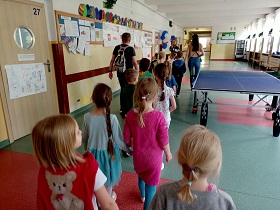 Dzieci idą w parach wraz z paniami spacerują po szkolnym korytarzu. Przechodzą obok stołu do gry w tenisa stołowego. 