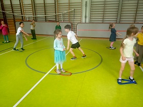 Dzieci biegają po sali gimnastycznej. Część z nich stoi stopami w szarfach. 