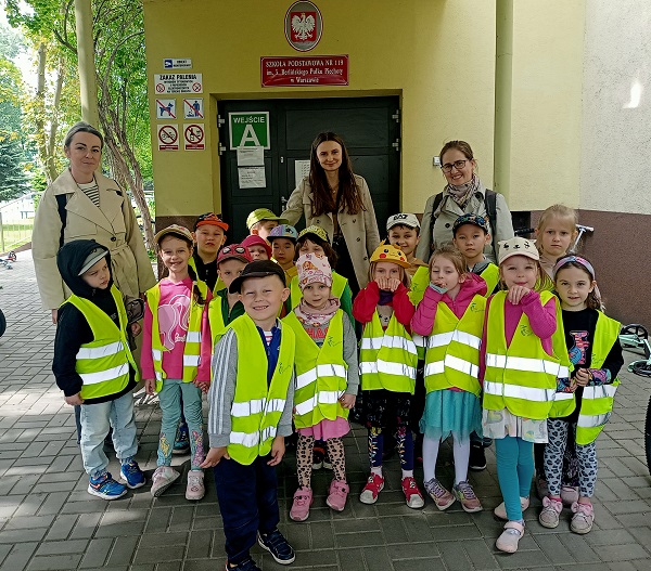 Dzieci ubrane w kamizelki odblaskowe wraz z trzema paniami stoją przed wejściem do szkoły podstawowej nr 119 w Warszawie.