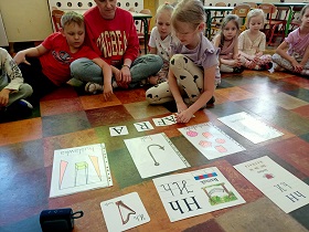 Dzieci siedzą w półkolu i obserwują dziewczynkę, która układa słowo harfa z kartoników z literami. Obok kartoników znajdują się ilustracje przedstawiające słowa na h. 