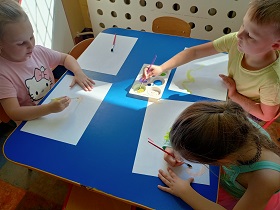 Dwie dziewczynki i chłopiec siedzą przy niebieskim stoliku i malują farbami po białych kartkach A3. Na stole znajduję się również paleta z farbami. 